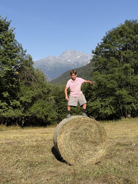 A young boy in a pink polo shirt and shorts stands balancing on a large hay bale in a sunny field